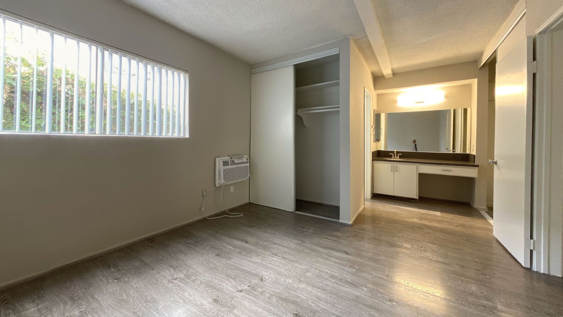 Empty apartment bedroom with closet, window with blinds, and bathroom entrance.