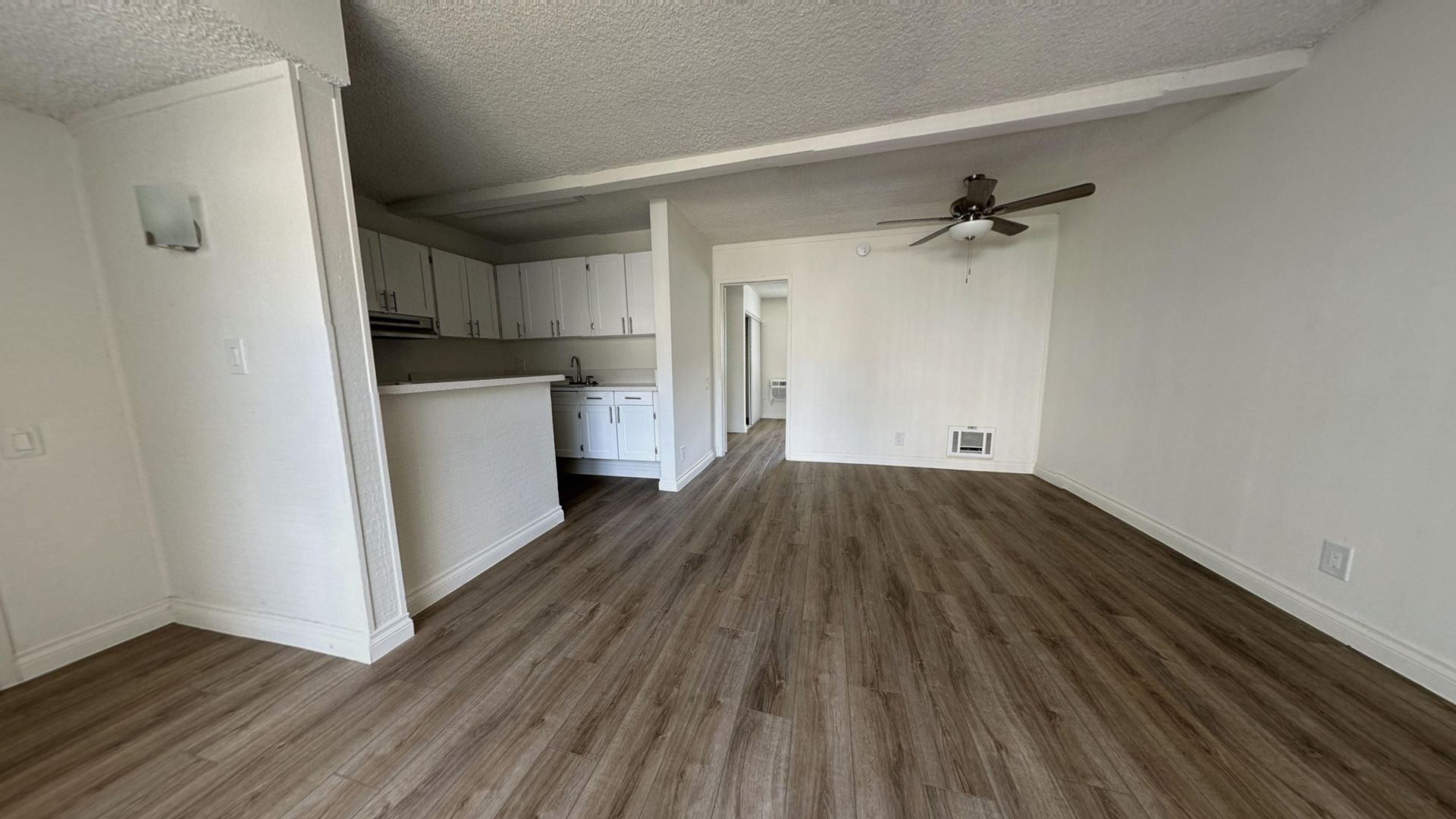 Empty apartment interior with wood-look flooring, white walls, ceiling fan, and kitchen visible in the background.