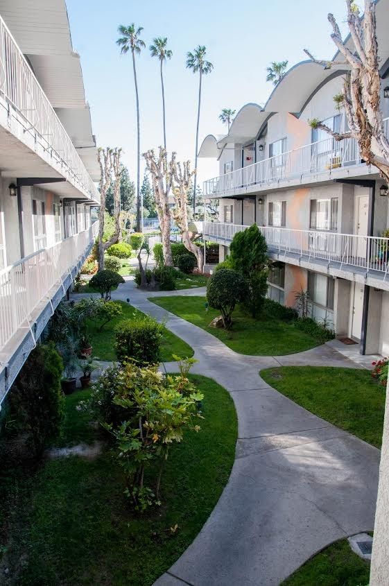 Apartment complex courtyard with walkways, greenery, and palm trees. Two-story buildings with balconies.