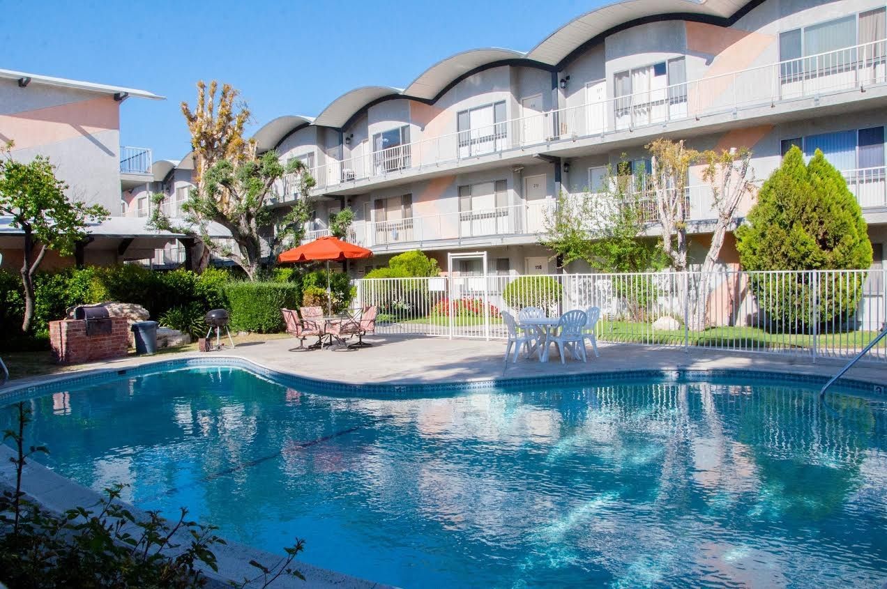 Pool in front of apartment building with arched roofs. Patio furniture and barbecue near the pool.