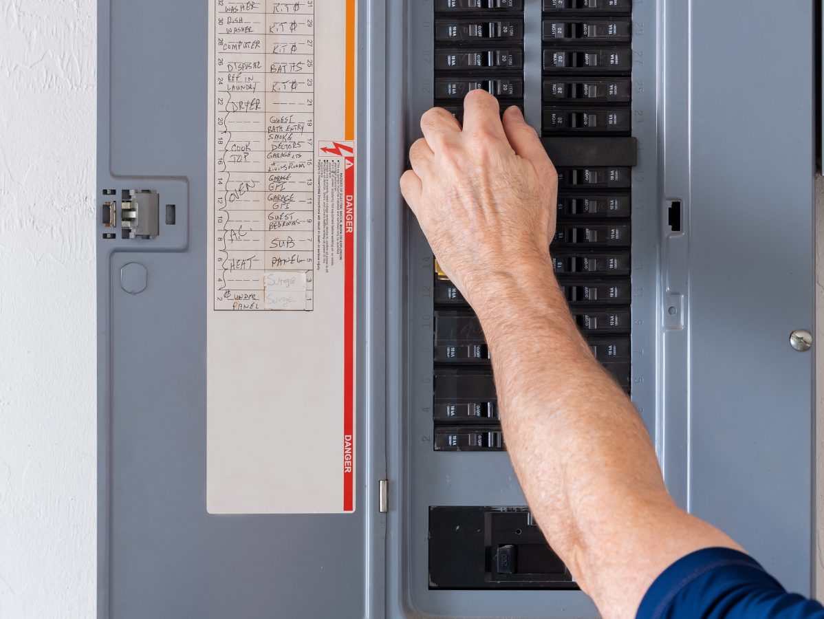Person's hand flipping a circuit breaker switch in a gray electrical panel, likely resolving a power issue.