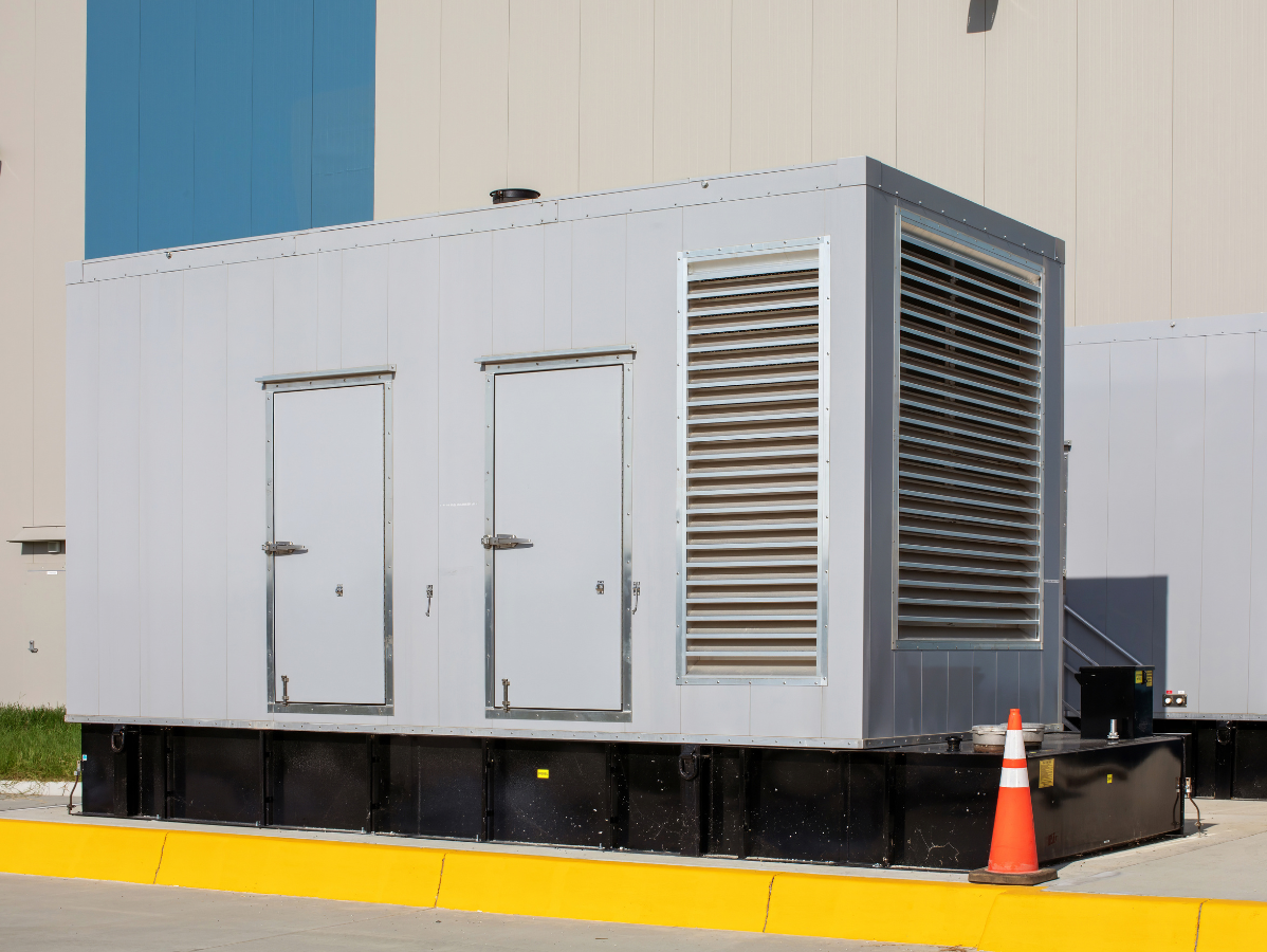 Generator in a gray enclosure, with doors and ventilation, sitting next to a building.