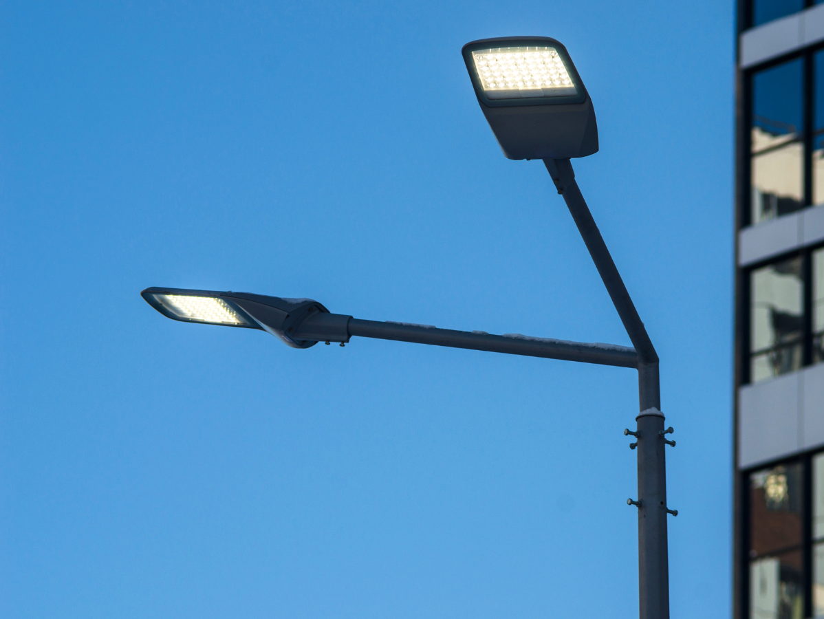 Streetlights with glowing rectangular LED fixtures against a blue sky and building.