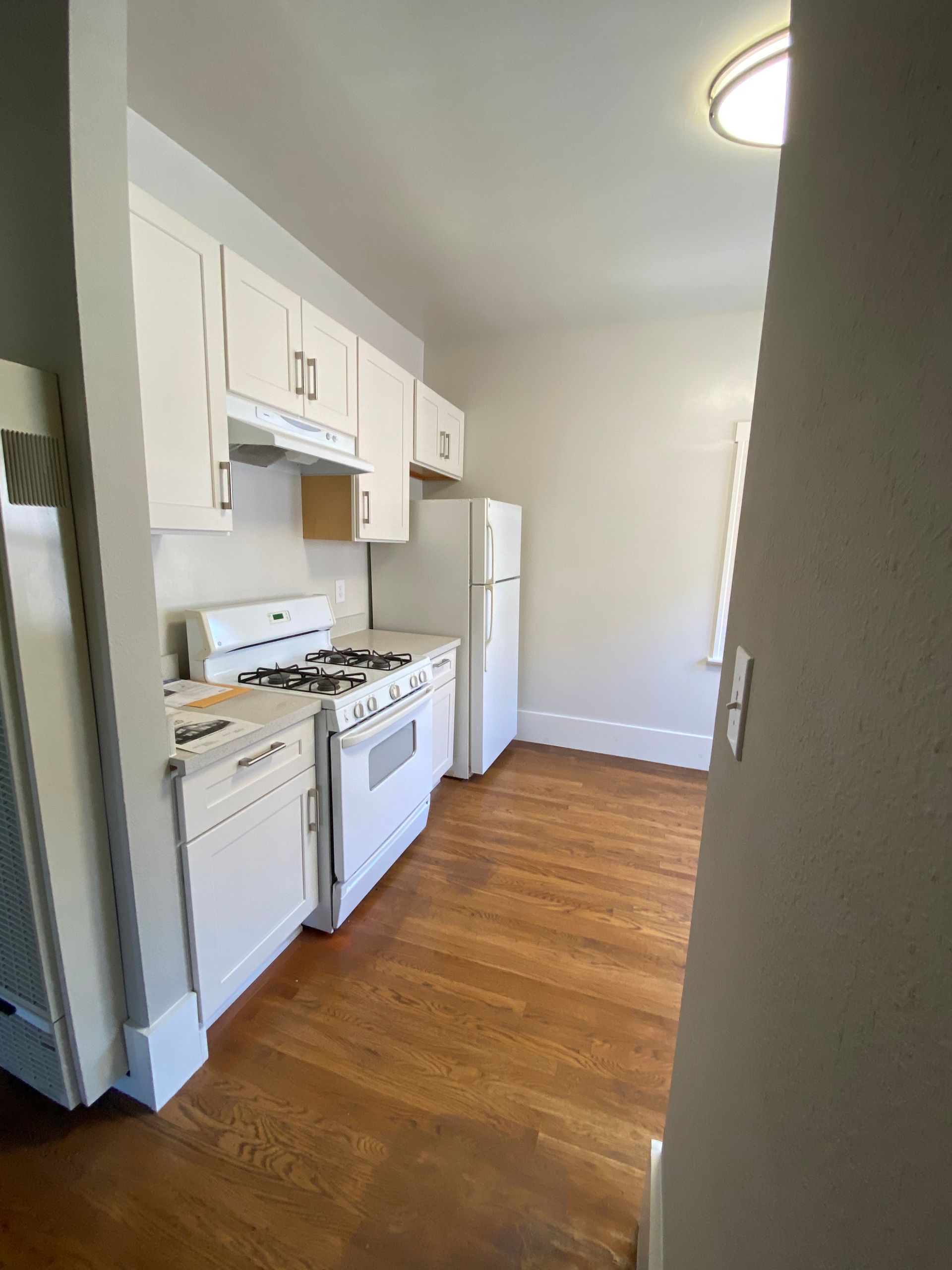 A kitchen with white cabinets , a stove , a refrigerator , and a wooden floor.