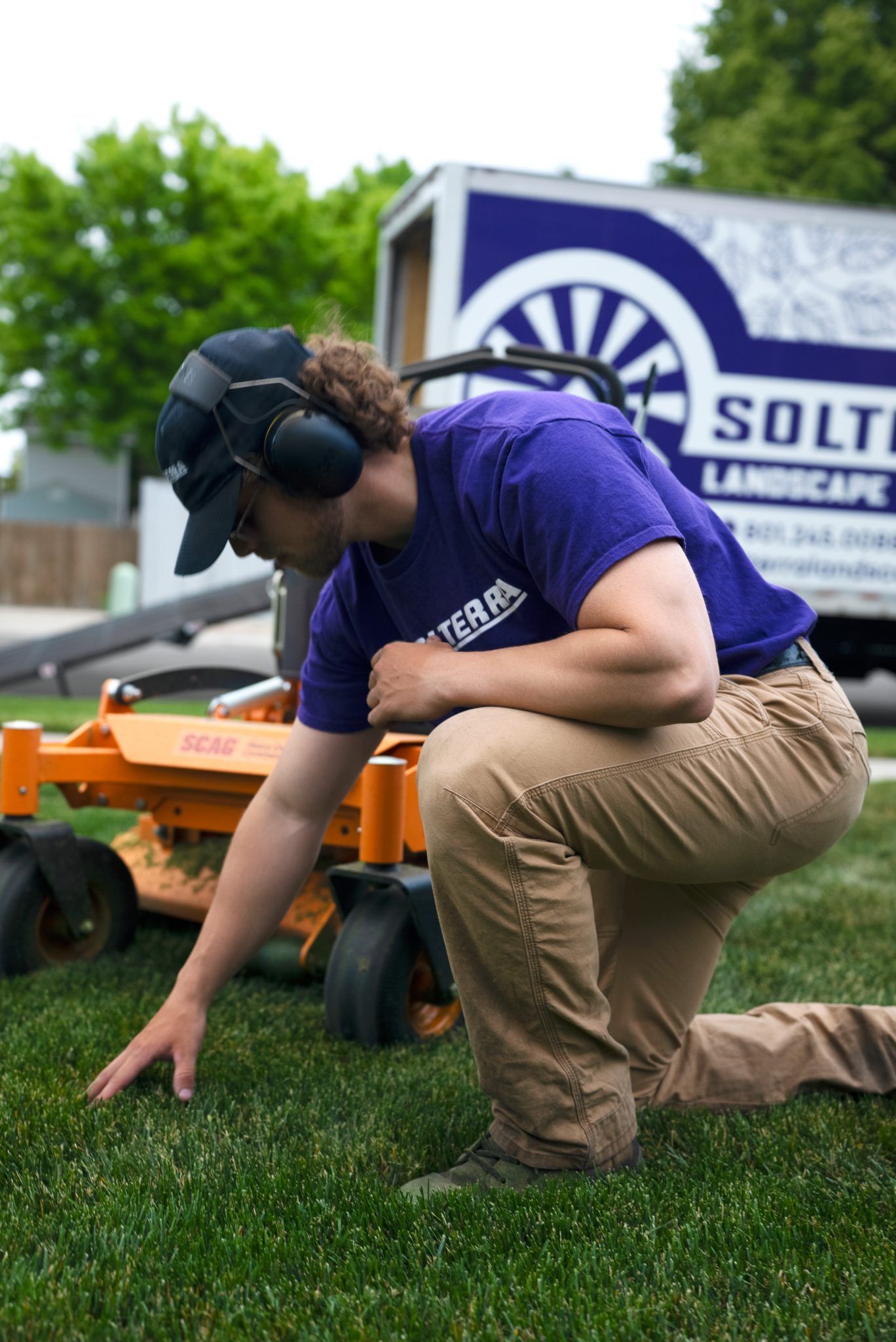 Landscaper kneels to inspect grass; orange mower, white truck, and blue sky background.