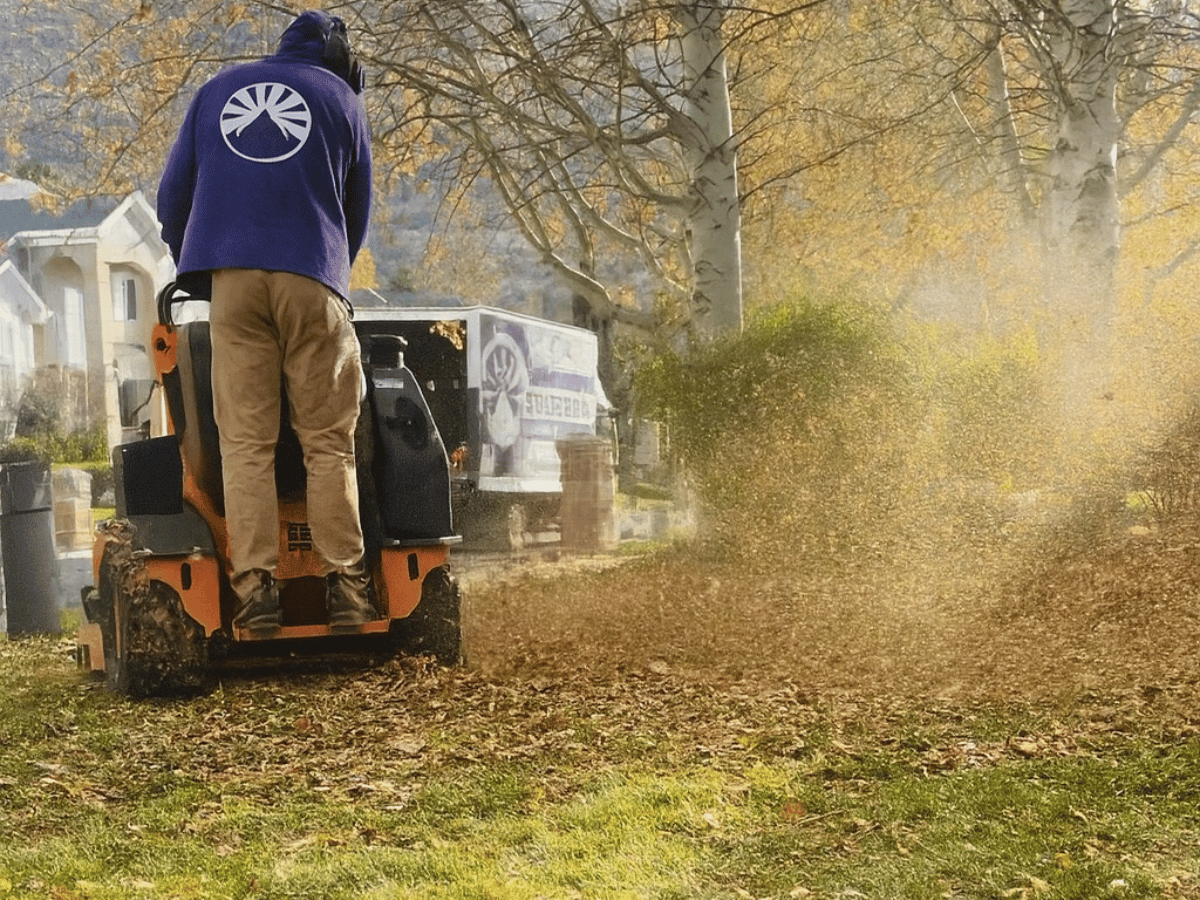 Man operating a leaf blower on an orange machine, blowing leaves on a lawn. A white truck is in the background.