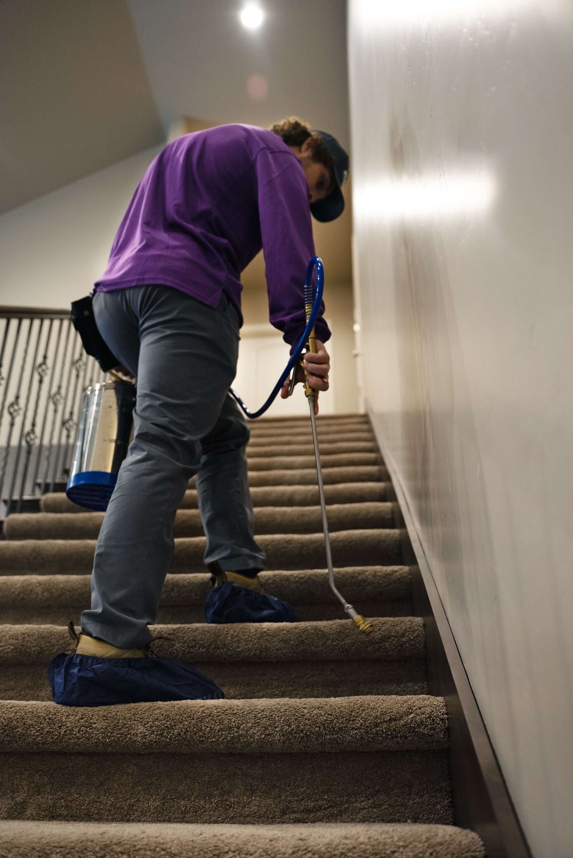 Person spraying stairs with pest control solution. They wear a purple shirt, blue jeans, and shoe covers.