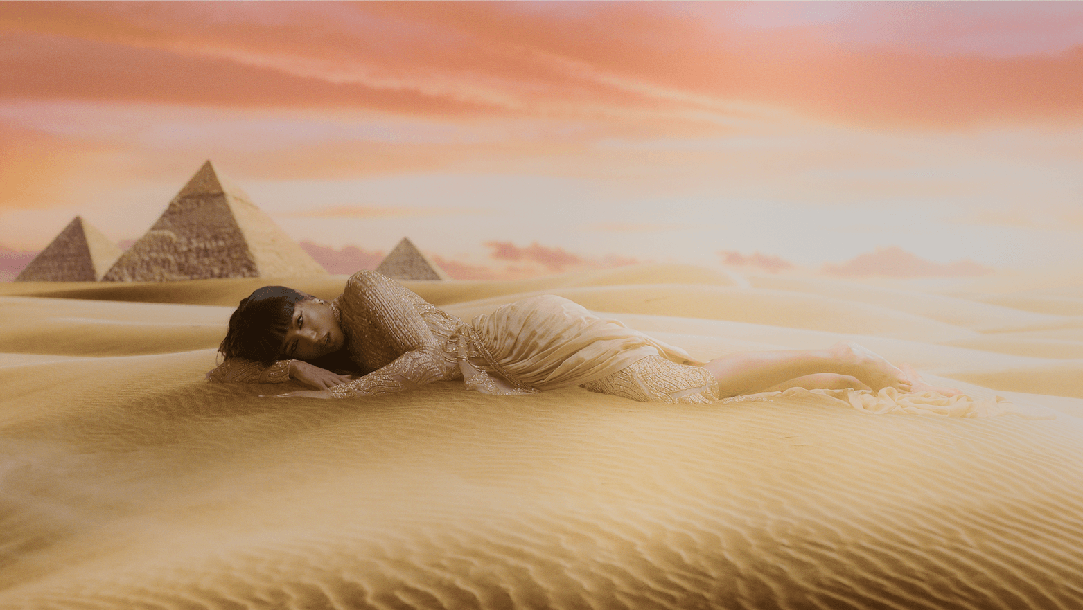 A woman is laying in the sand in the desert with pyramids in the background.