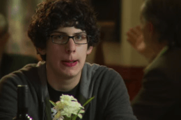 A young man wearing glasses is sitting at a table with a bottle of wine