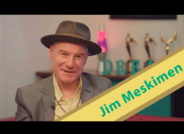 A man wearing a hat and a suit is next to a sign that says jim meskimen