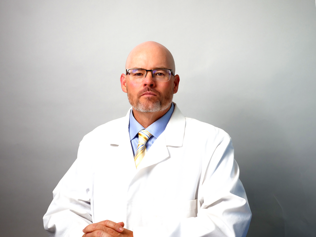 A man in a white coat and tie is sitting with his hands folded. PODIATRIST