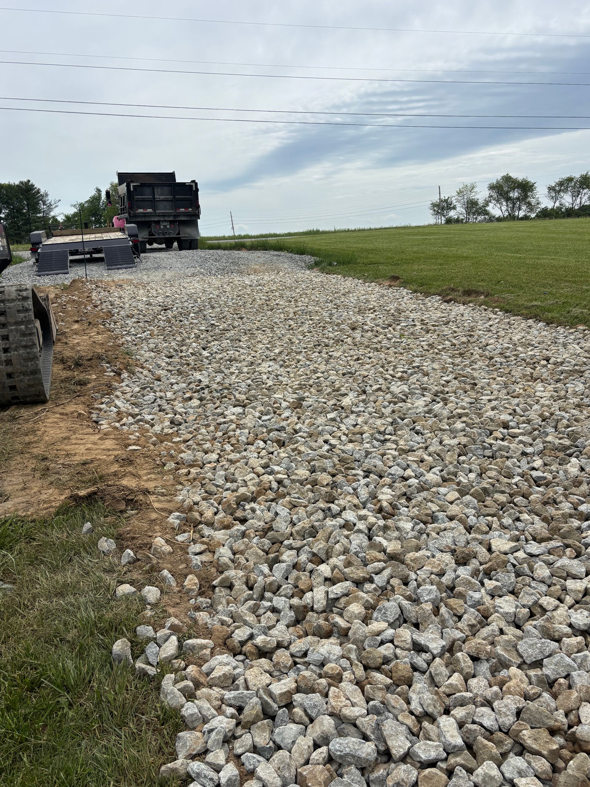 Gravel road under construction beside a green field, with a dump truck and paving equipment ahead