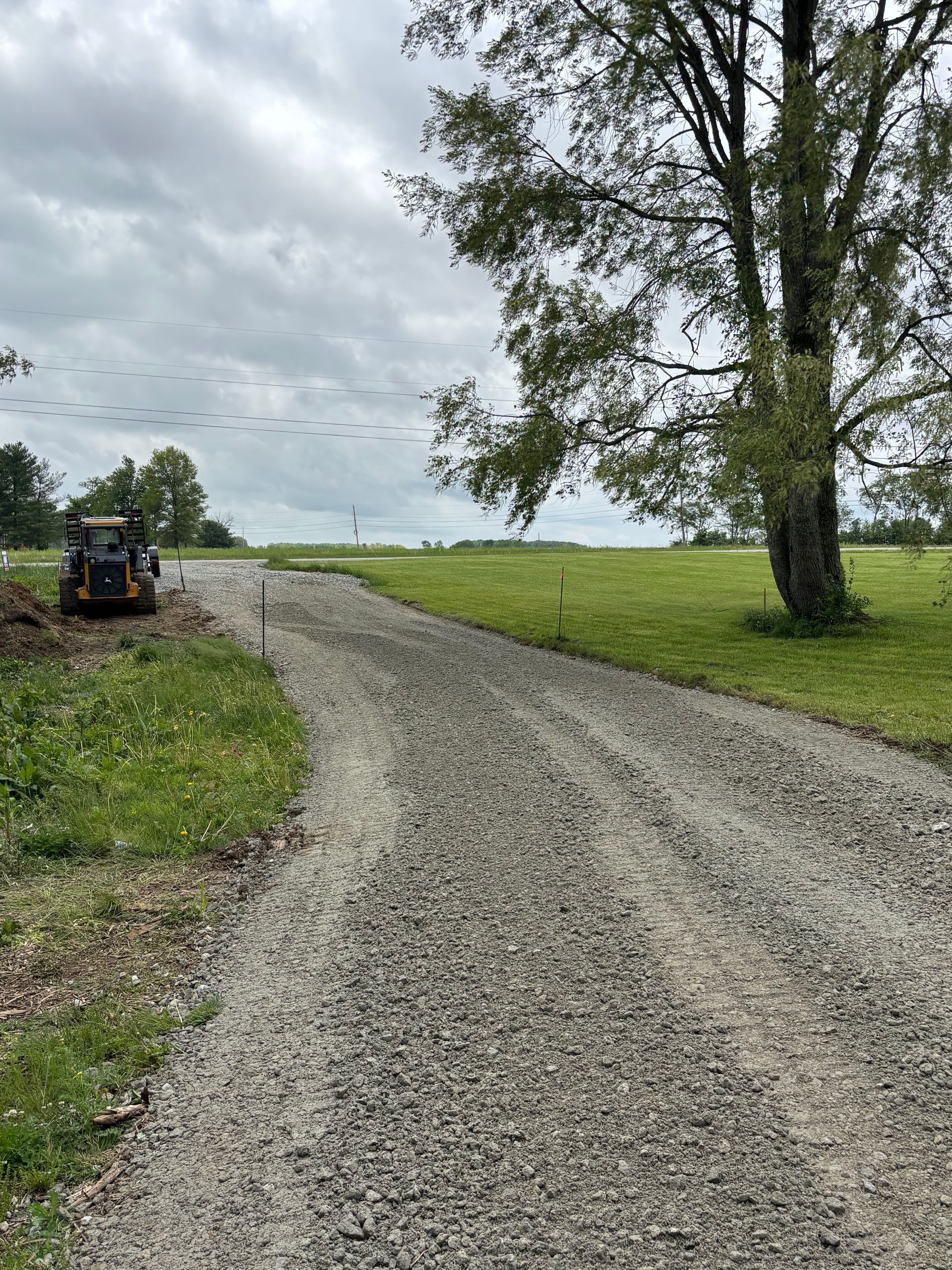 Gravel road curving past a green field and tall tree under cloudy sky, with a tractor on the left.
