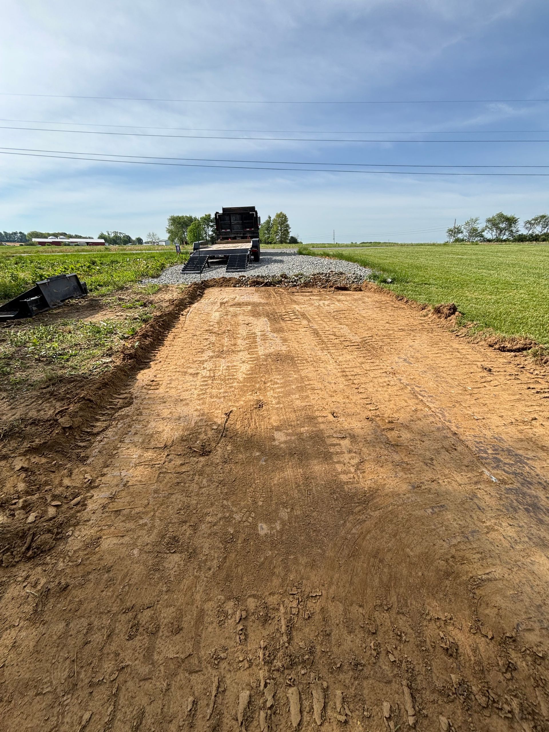 Dirt road under construction leading to a gravel section in open farmland, with a truck in the distance.
