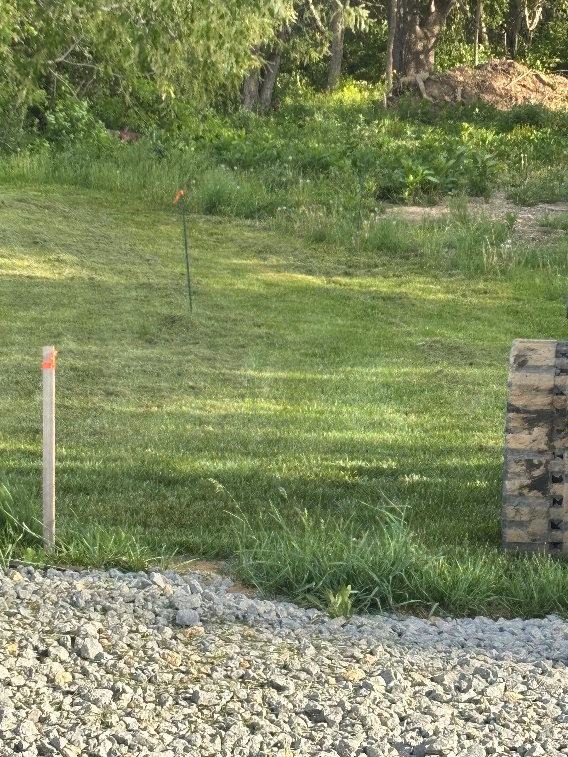 Grassy yard with gravel foreground, a survey stake, and a stone retaining wall section.