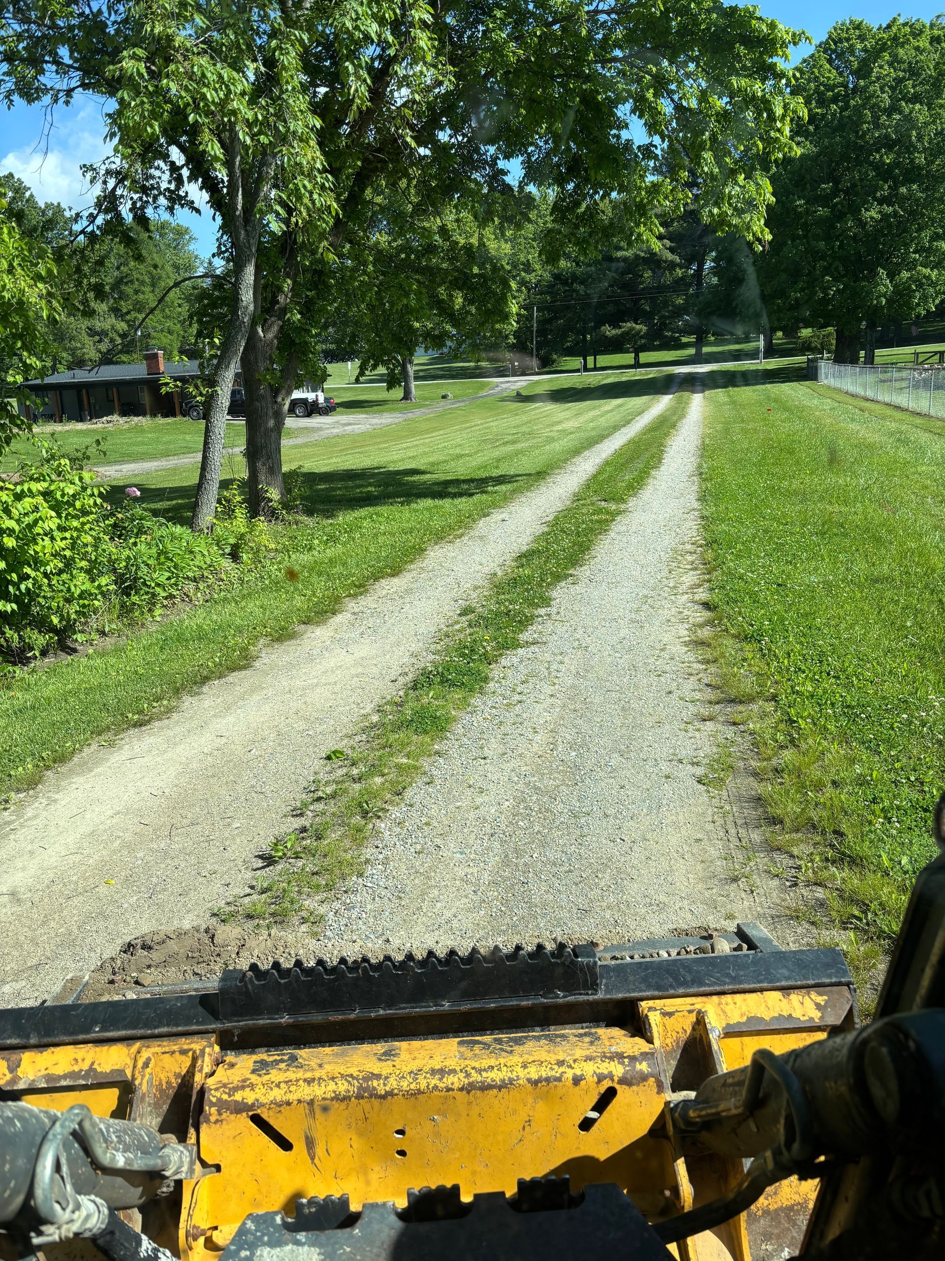 View from a yellow vehicle on a grassy gravel driveway lined with trees and a house in the distance