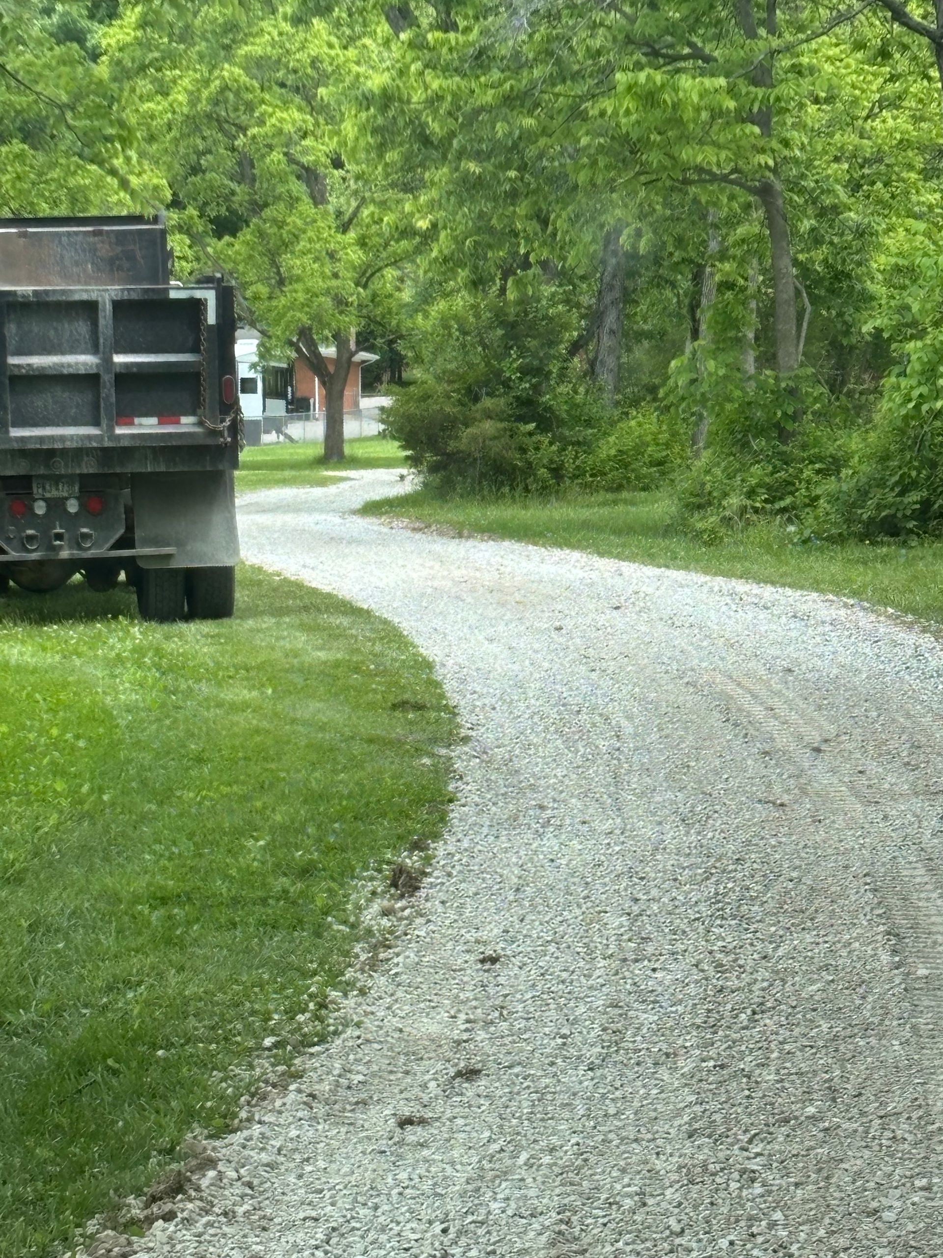 Gravel driveway curving through a green wooded area, with a dump truck parked at left.