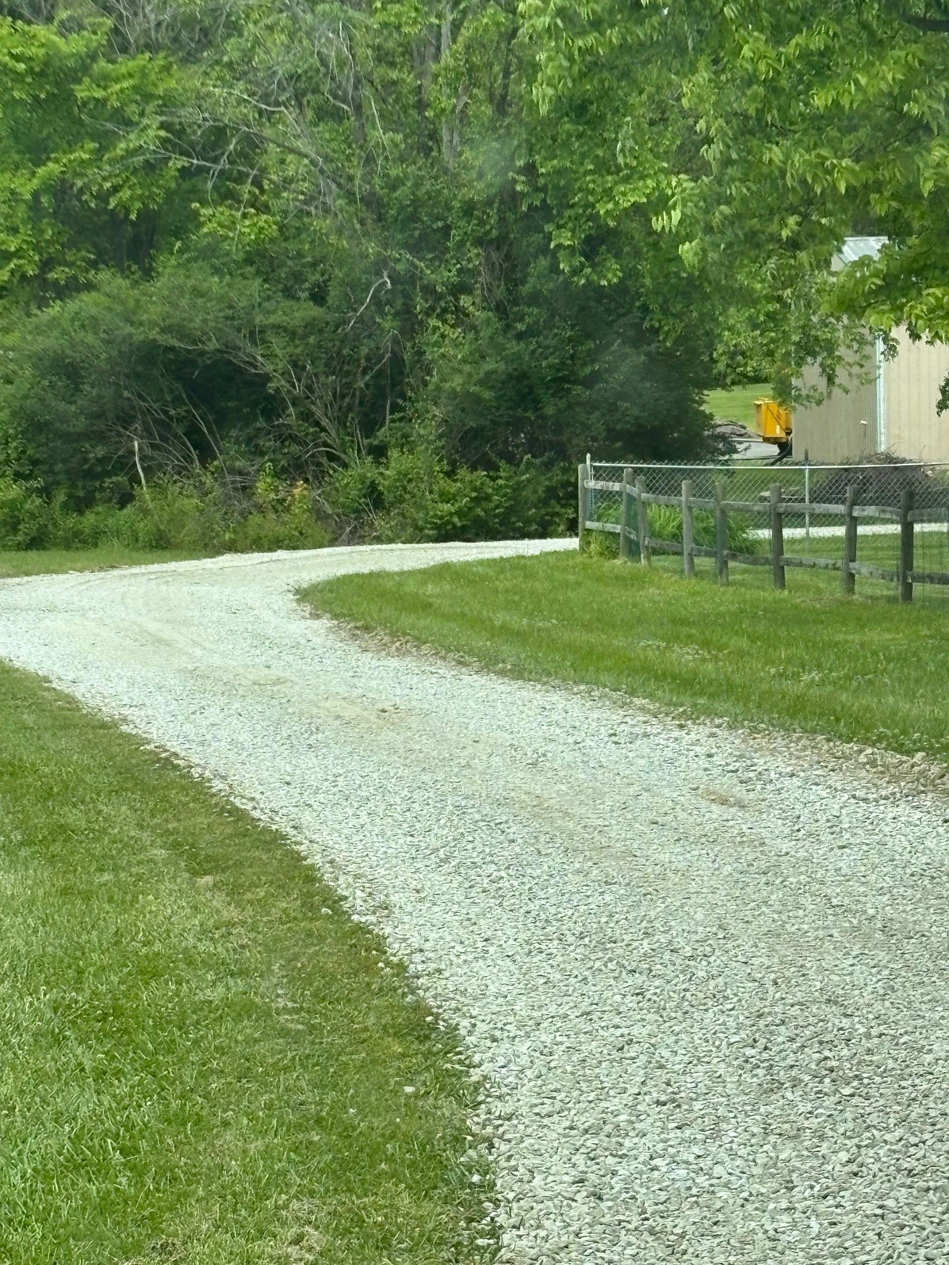 Curved gravel driveway through green trees beside a fenced yard and white building