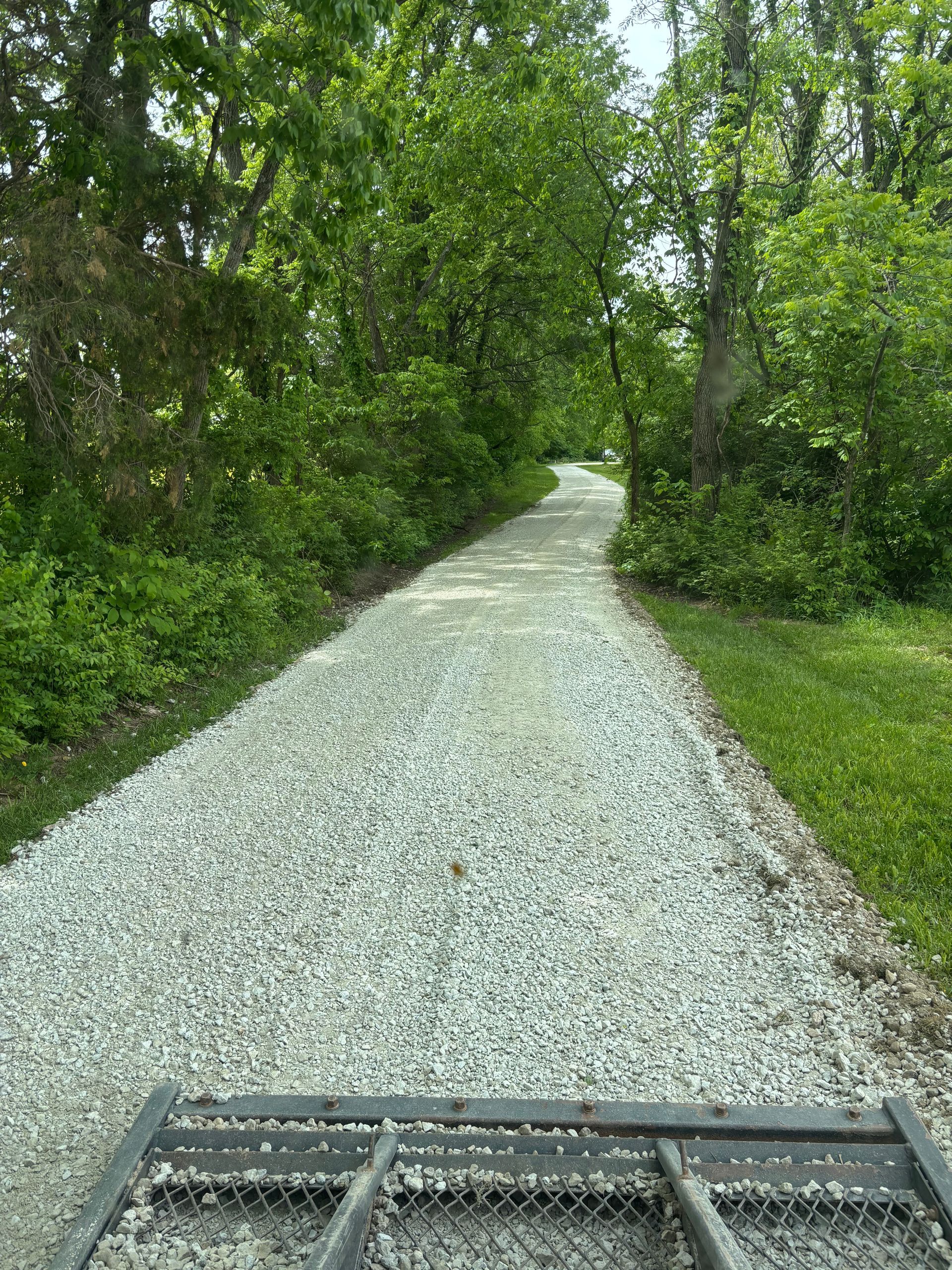 Gravel road through dense green trees, seen from a vehicle’s front grille.