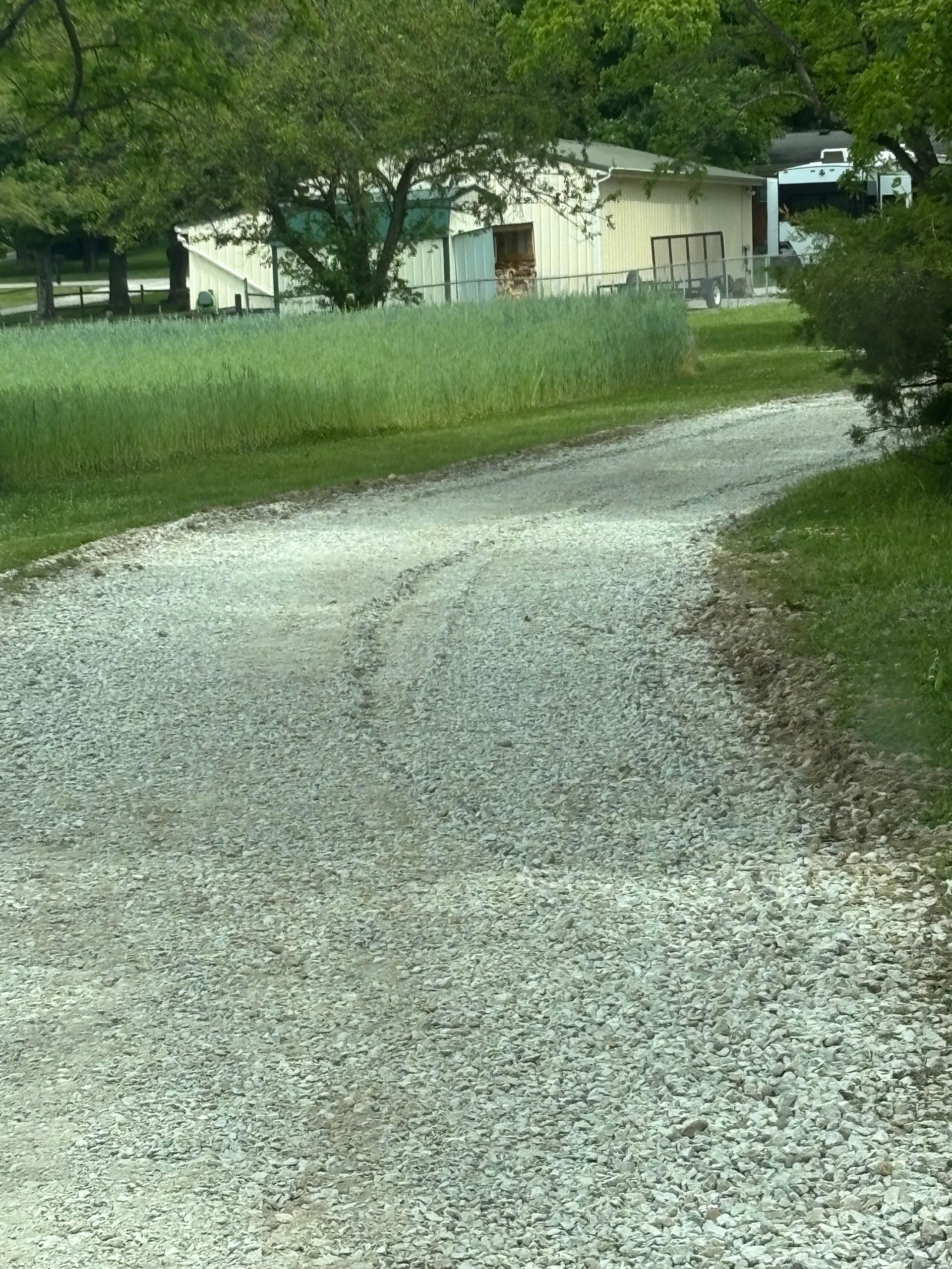 Gravel driveway curving past a grassy field toward a small beige building with trees nearby