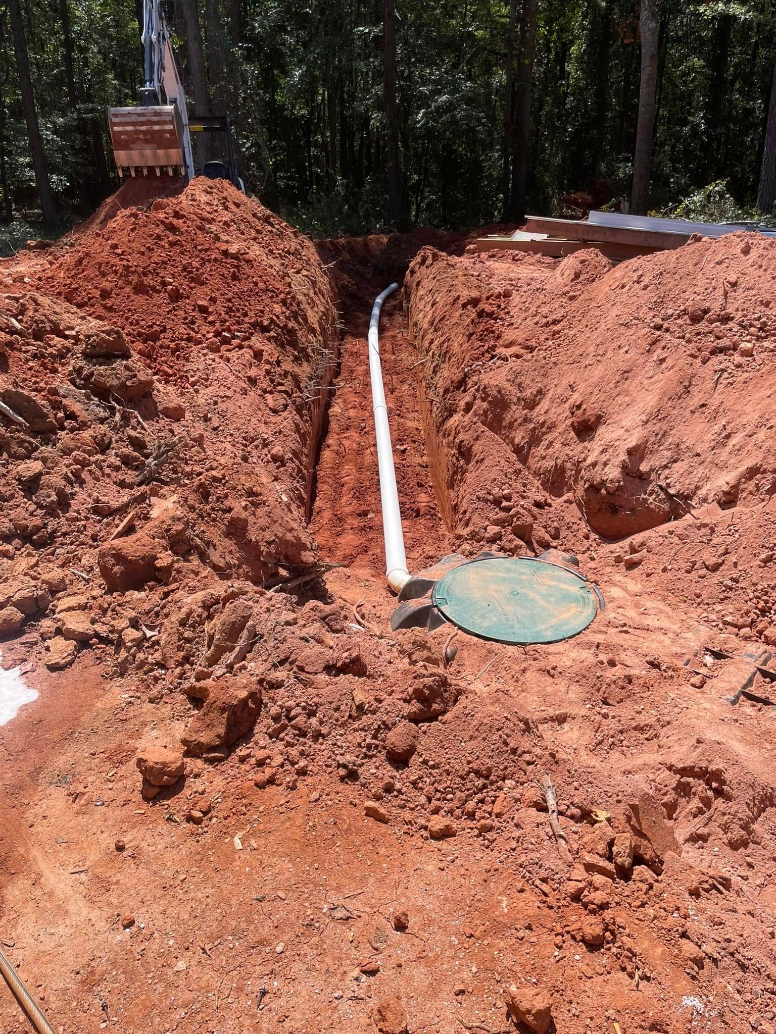 A trench dug in red soil with white pipe and a green septic tank lid.