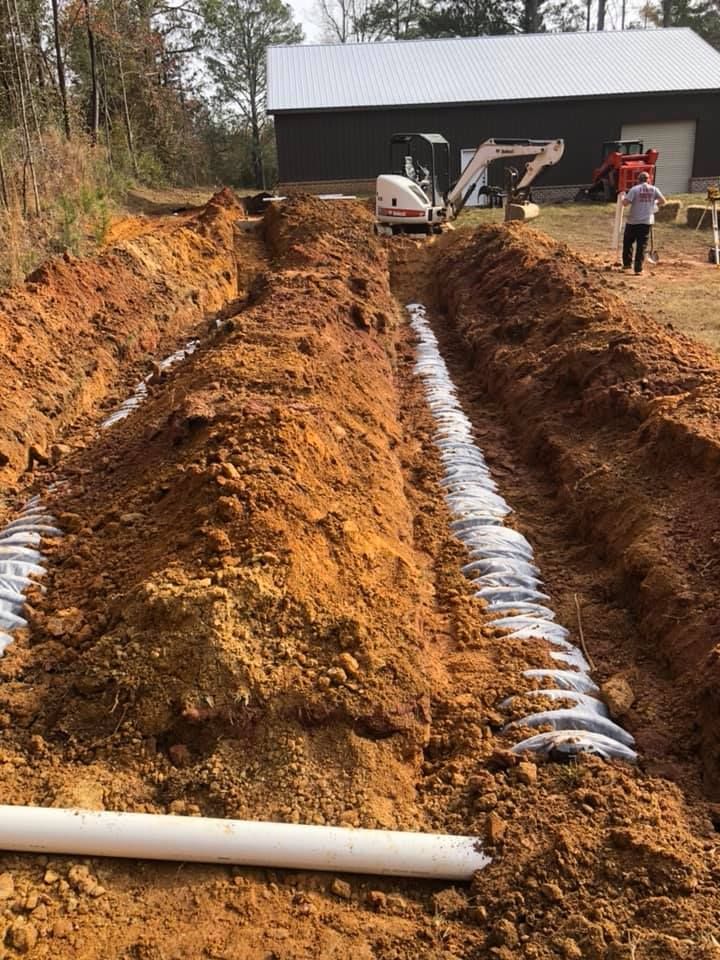 Trenches dug for a septic system installation, with white pipes and building in background.
