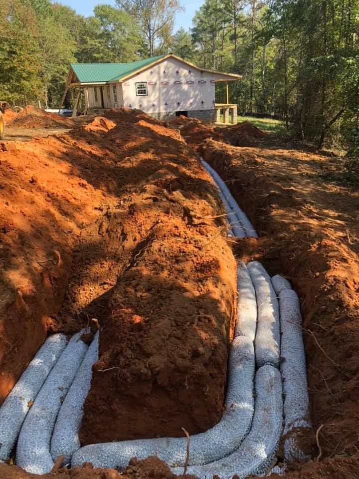 Trench with white septic system components installed. House under construction in the background. Sunny, outdoor setting.
