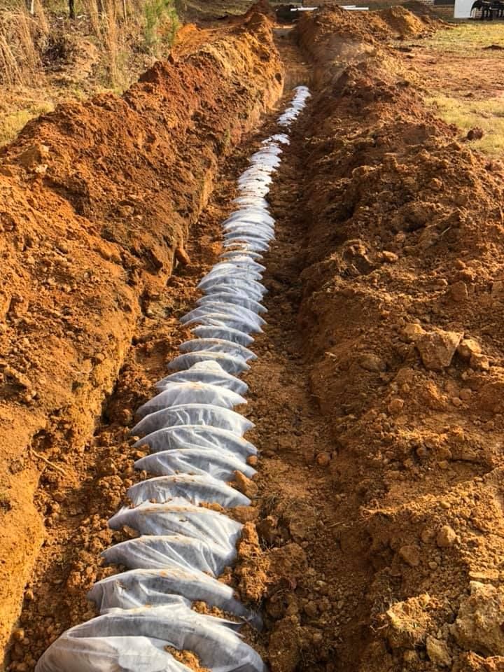 A long trench lined with white fabric and circular pipe, set in brown dirt, outdoors.