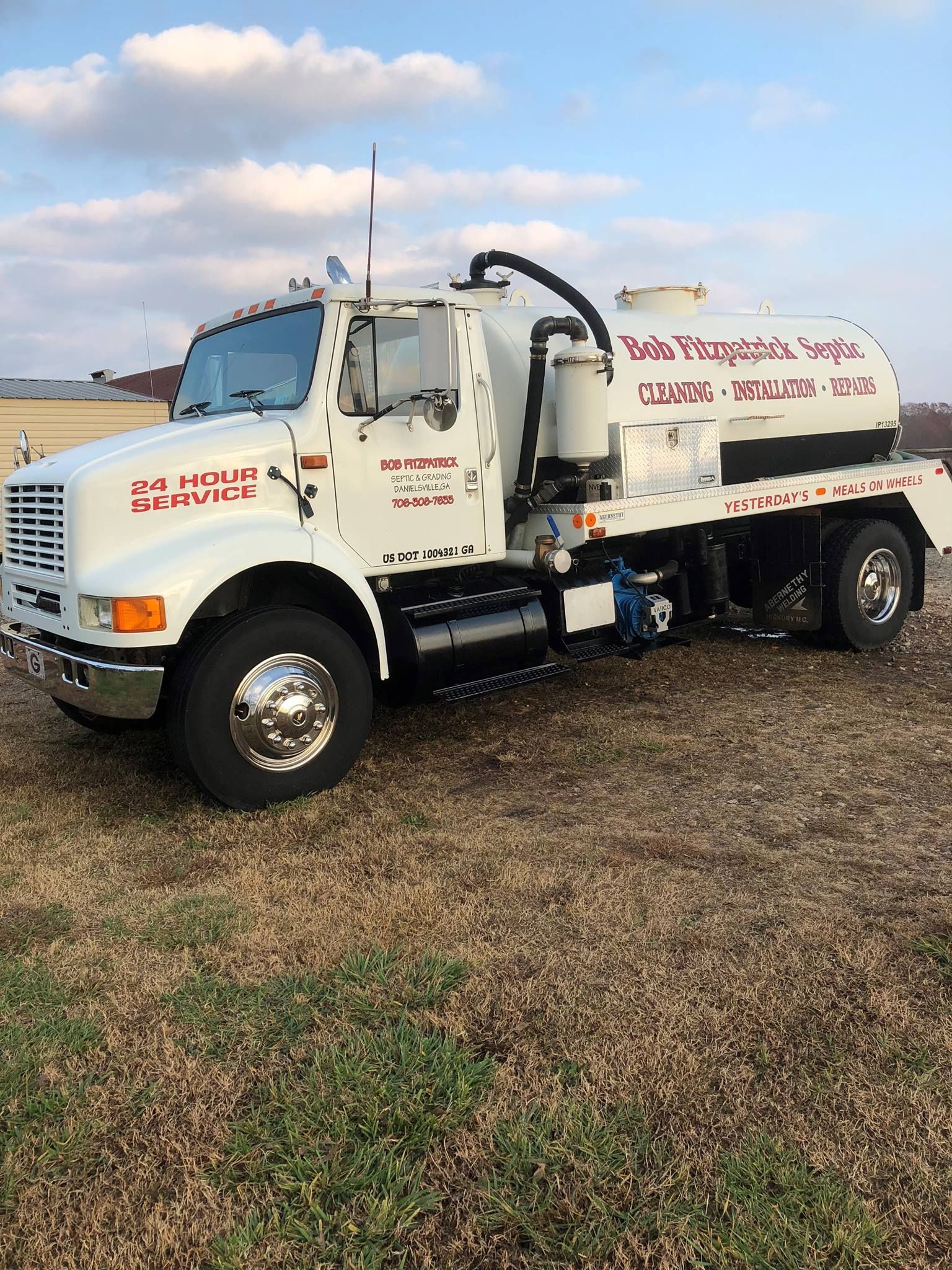 White septic service truck parked on grassy field. 24-hour service written on the door.