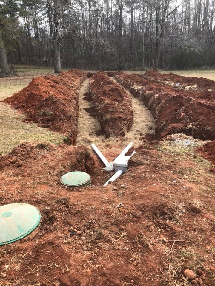 Septic system installation: trenches dug in red soil, pipes connected, green tank lids visible, trees in the background.