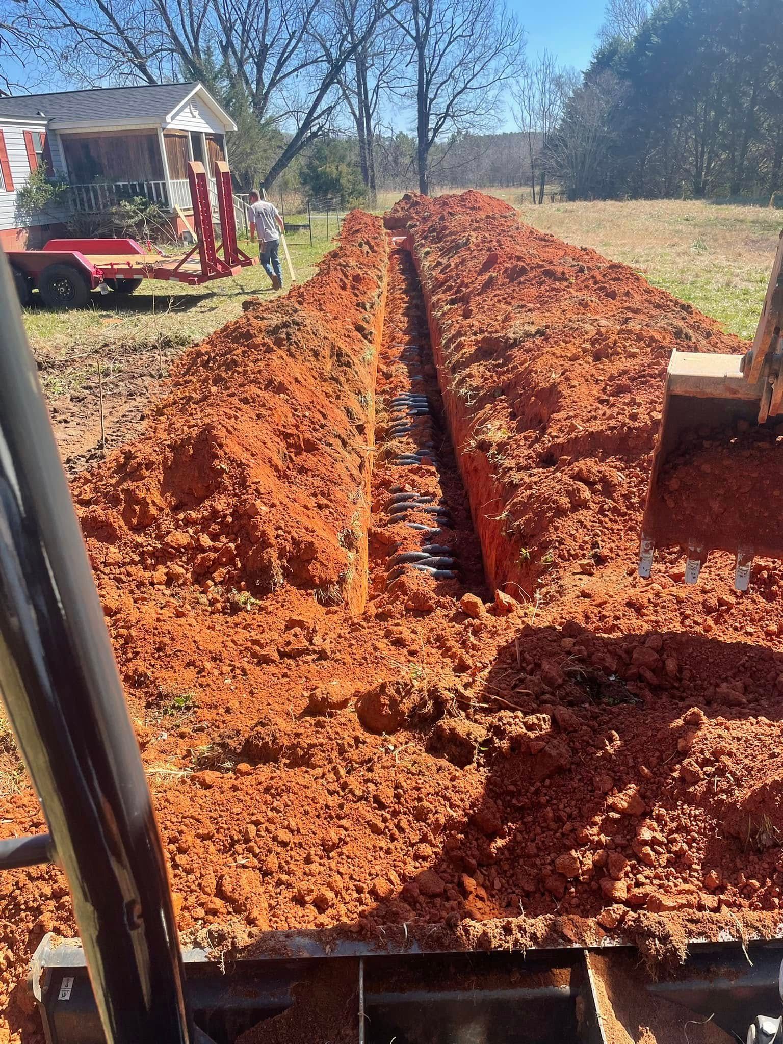 A backhoe digging a long trench in red soil, a person walking in the background.