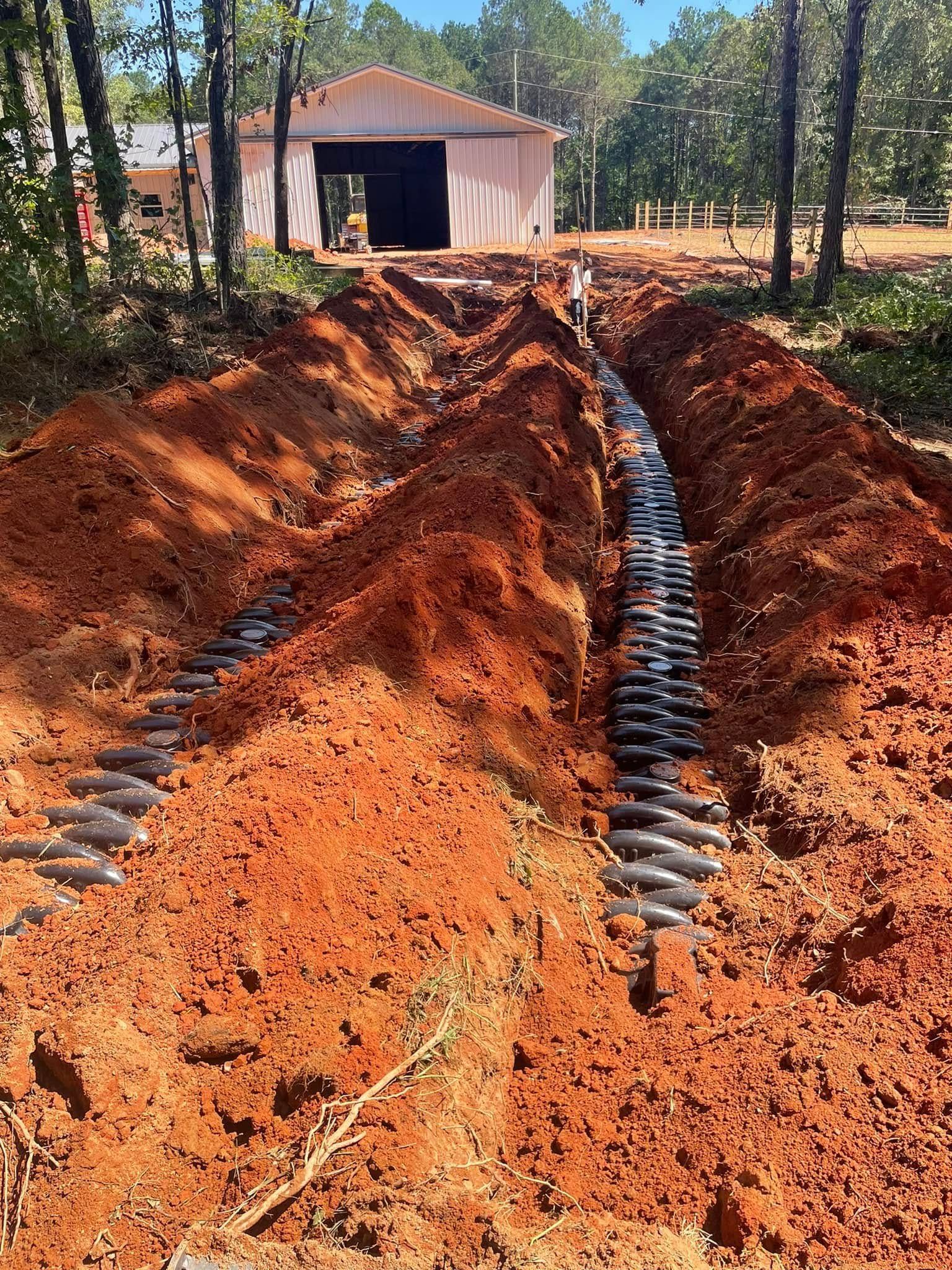 Trench dug in red soil with black pipes, likely part of a septic system, leading to a building.