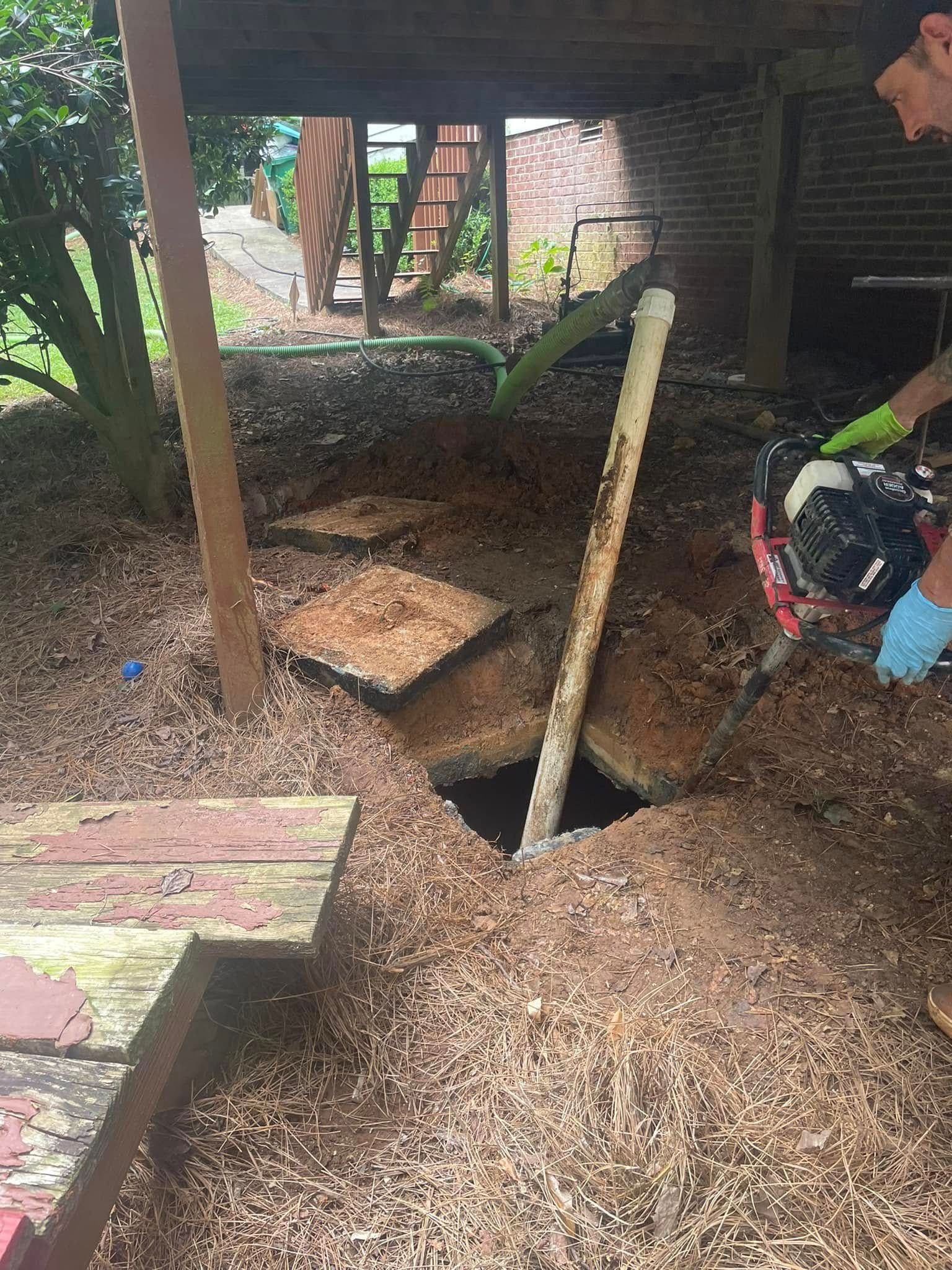 Hole dug under deck with exposed pipe. Someone wearing gloves operates equipment. Brown, natural setting.