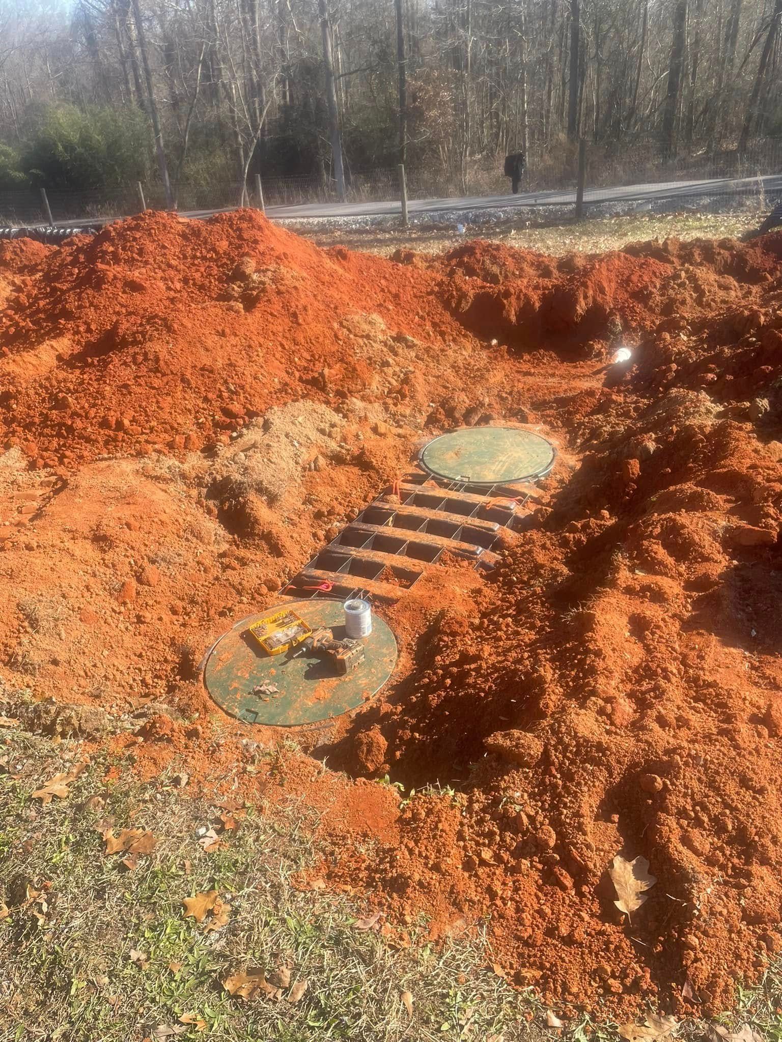 Excavated septic system components in red clay, with visible access lids, outdoors.