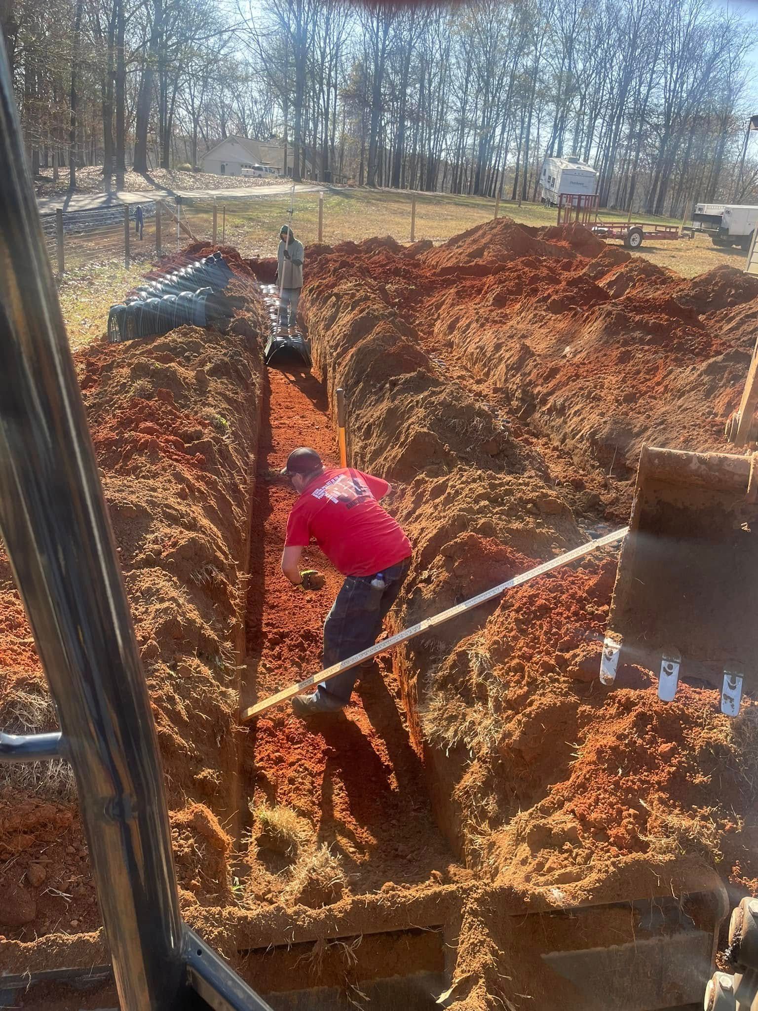 Man in red shirt working in a trench. Brown soil, white pipe, trees and a clear day.