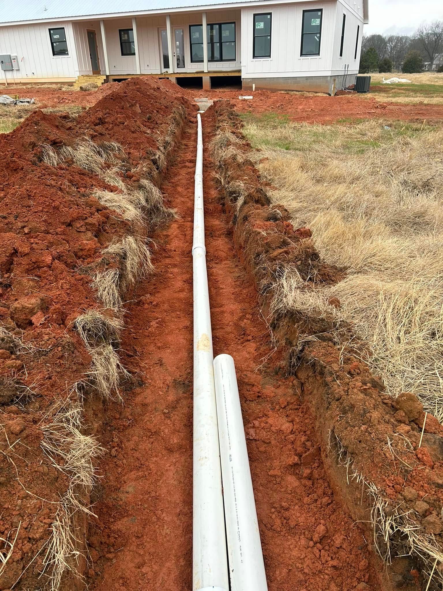 Trench with white pipes in front of a house under construction; red dirt and grass.
