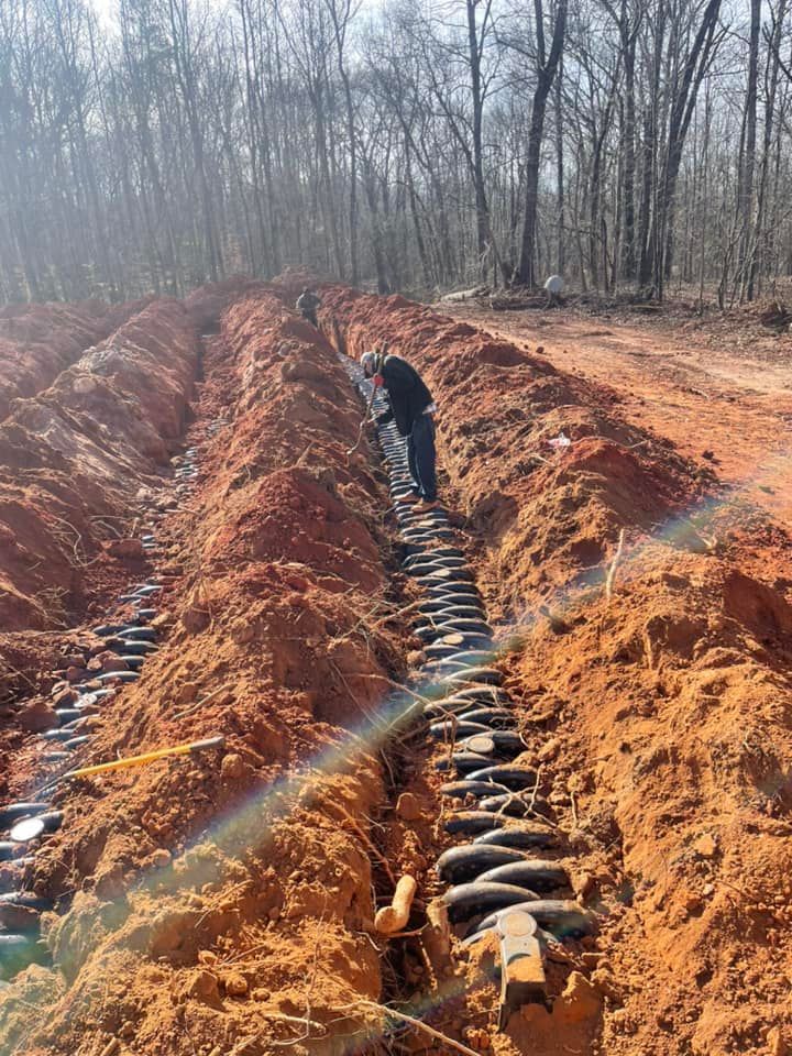 Person inspecting drainage system in a dirt trench, red clay soil, forest background, sunny day.