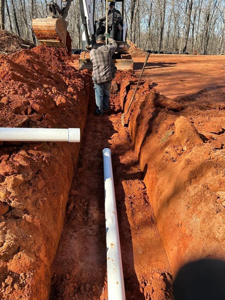 Man installing white PVC pipes in a trench dug by an excavator on a sunny day.