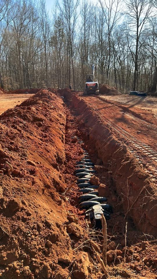 A trench filled with black septic system components in red dirt, with an excavator in the background.