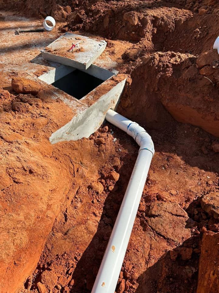 White PVC pipe curving toward an open underground access point in red soil. A shovel rests on the pipe.