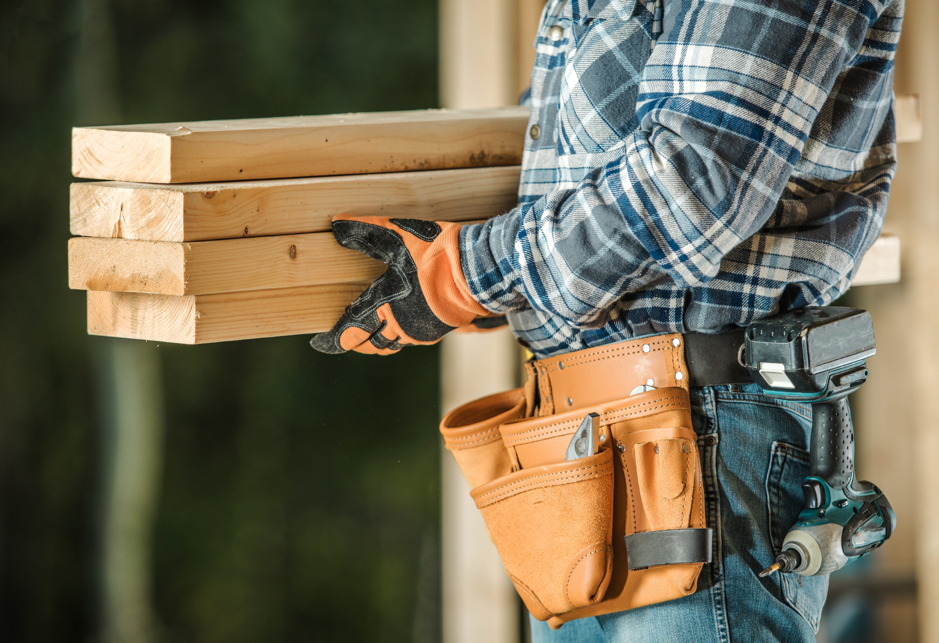 Carpenter carrying wooden boards at a construction site, wearing a tool belt and gloves.
