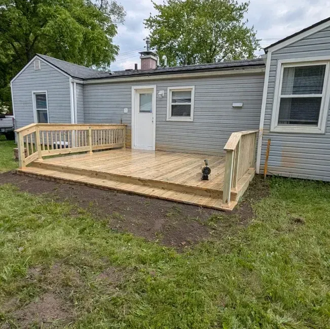 Wooden deck with railings attached to a light gray house; backyard setting with grass and dirt.