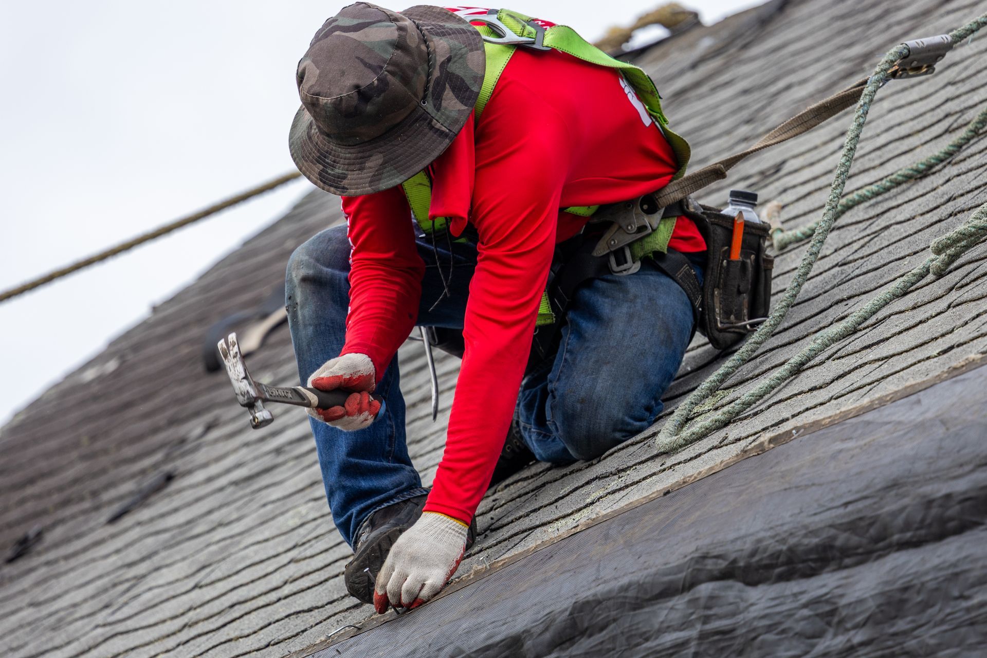 A man is kneeling on a roof using a hammer.