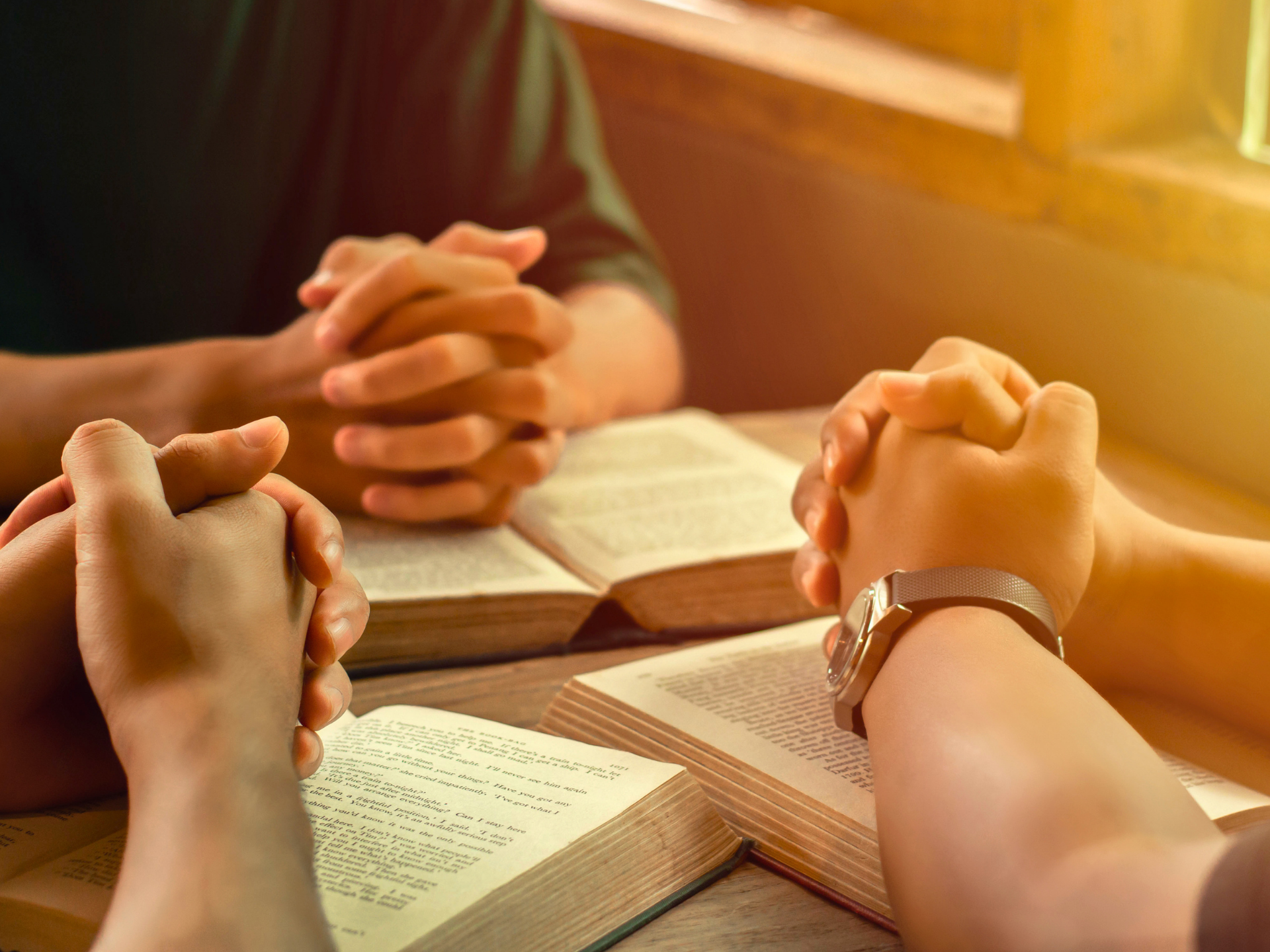 People with clasped hands over open books in a warm-lit study group setting