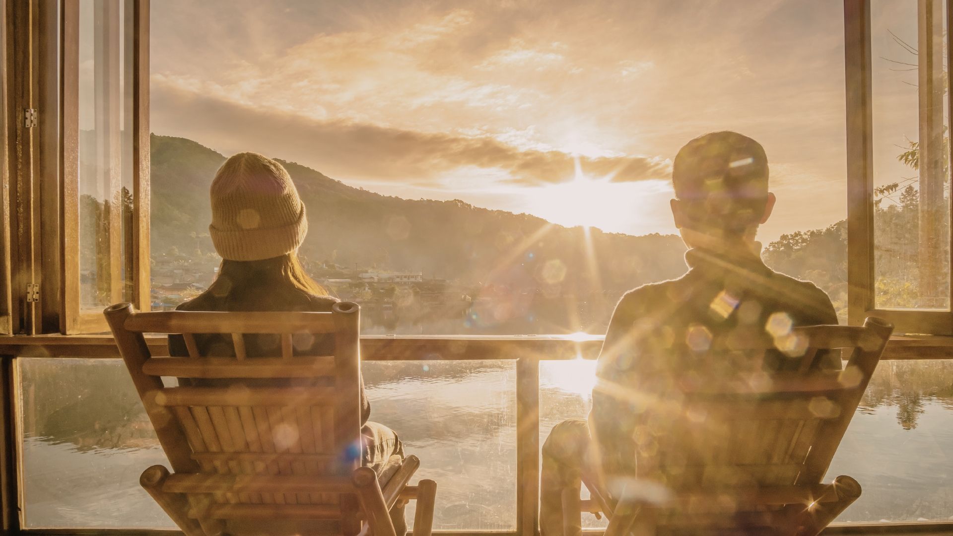 Two silhouetted people in chairs watching a sunset through a large window.