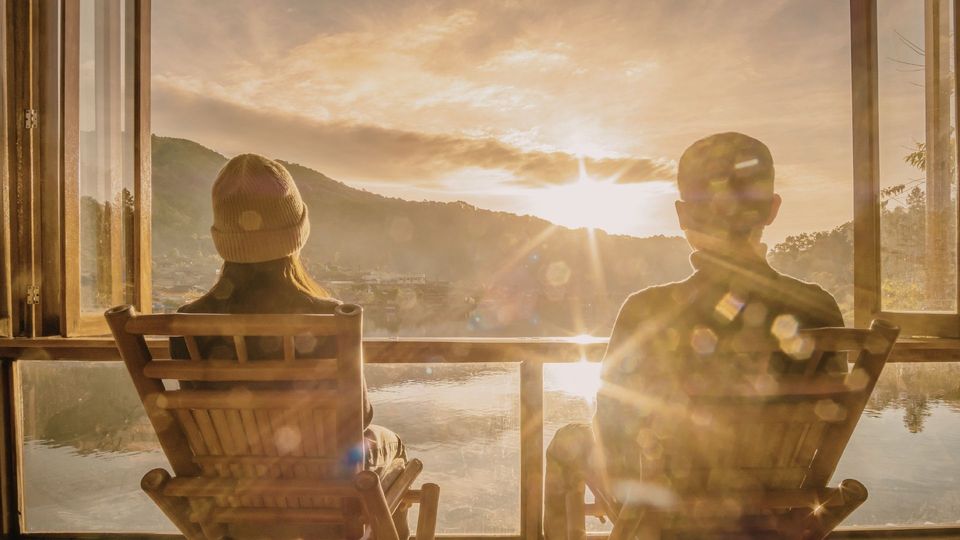 Two silhouetted people in chairs watching a sunset through a large window.