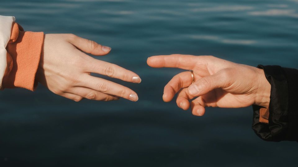 Two hands reaching toward each other over dark water, one from the left in orange sleeve, one from the right in black.