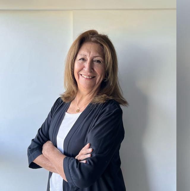 Una mujer con una chaqueta negra y una camisa blanca está de pie con los brazos cruzados y sonriendo.