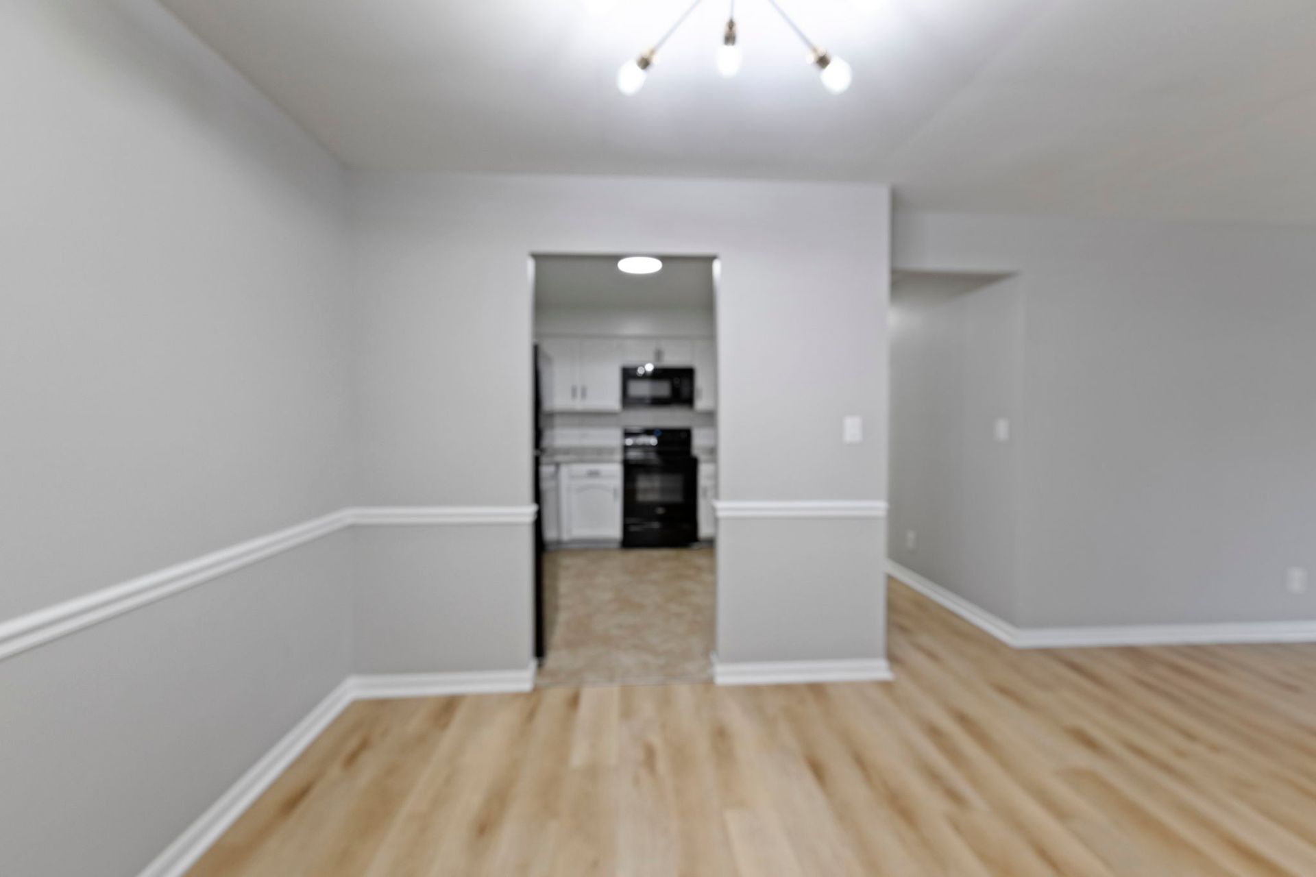 Empty dining room with light gray walls, white trim, and wood-look floors, leading to a black-and-white kitchen.