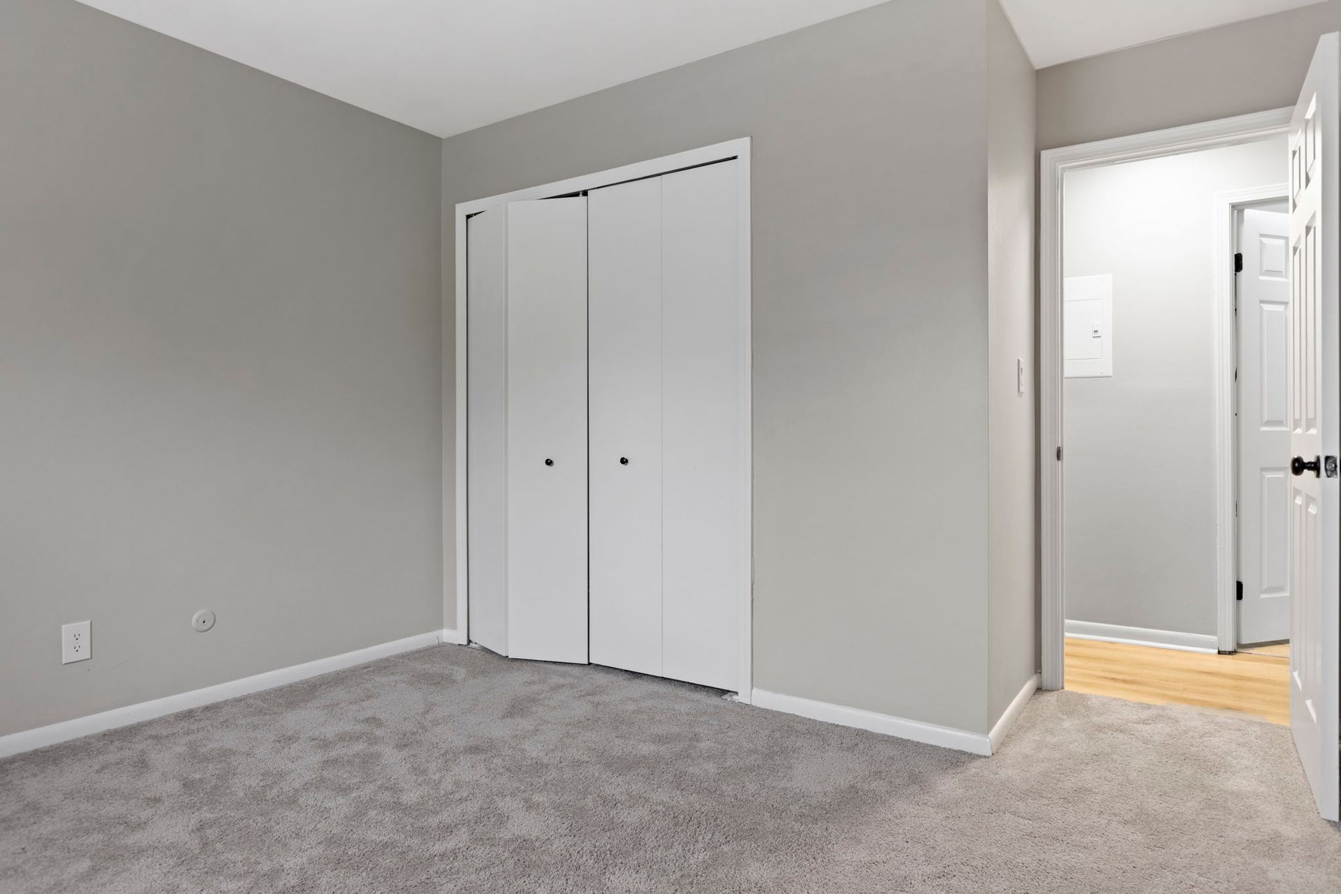 Empty bedroom with gray walls, carpet, and white closet doors. Doorway to another room on the right.